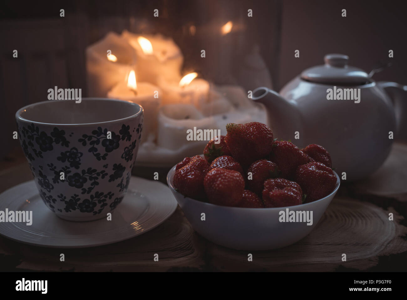 Teiera, una tazza e una ciotola di fragole contro candele accese Foto Stock