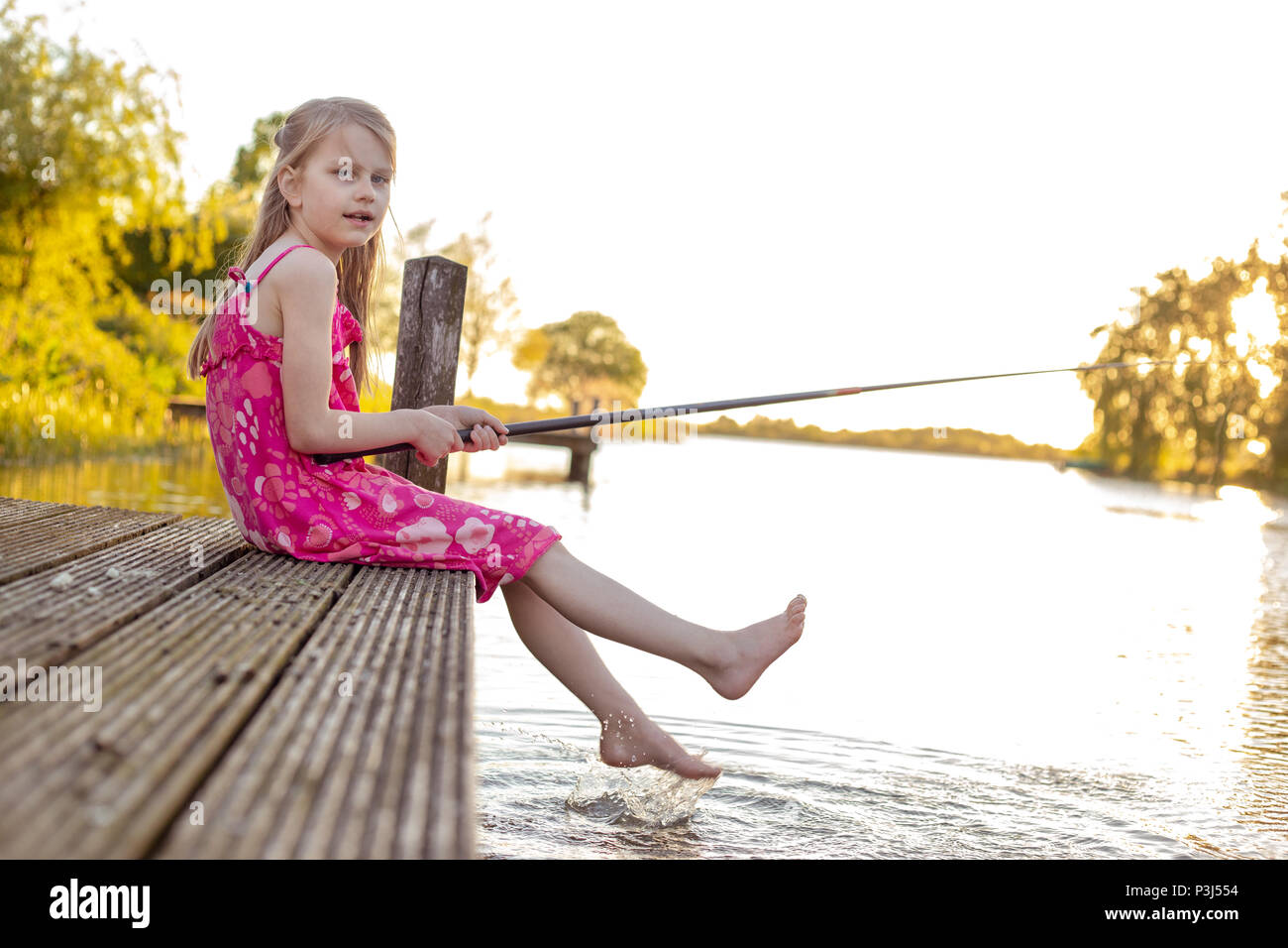 Una bambina di 7 anni seduto con la sua canna da pesca su un molo da un  lago, in attesa di un pesce a mordere. Lei si guarda nella telecamera e  spruzzi