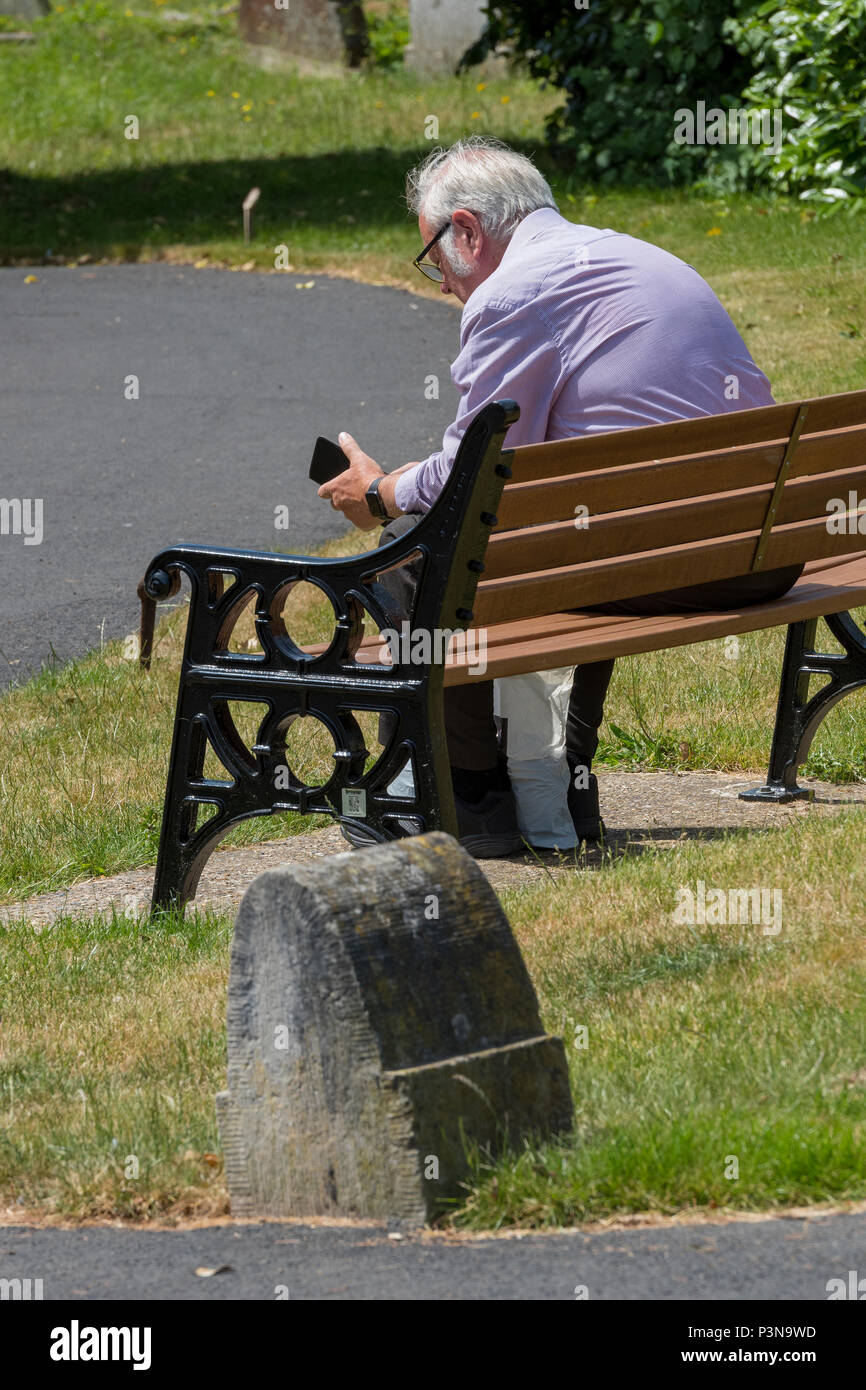 Un vecchio o anziano uomo silver surfer seduta su una panchina in un parco utilizzando uno smartphone o un dispositivo portatile per e-mail e leggere i messaggi di posta elettronica. generazione di anziani. Foto Stock