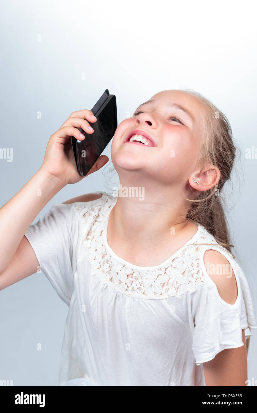 Una bella giovane ragazza caucasica (10 anni) in bianco estate shirt ridendo ad alta voce mentre si tiene un nero telefono cellulare, sentimenti positivi su una luce Foto Stock