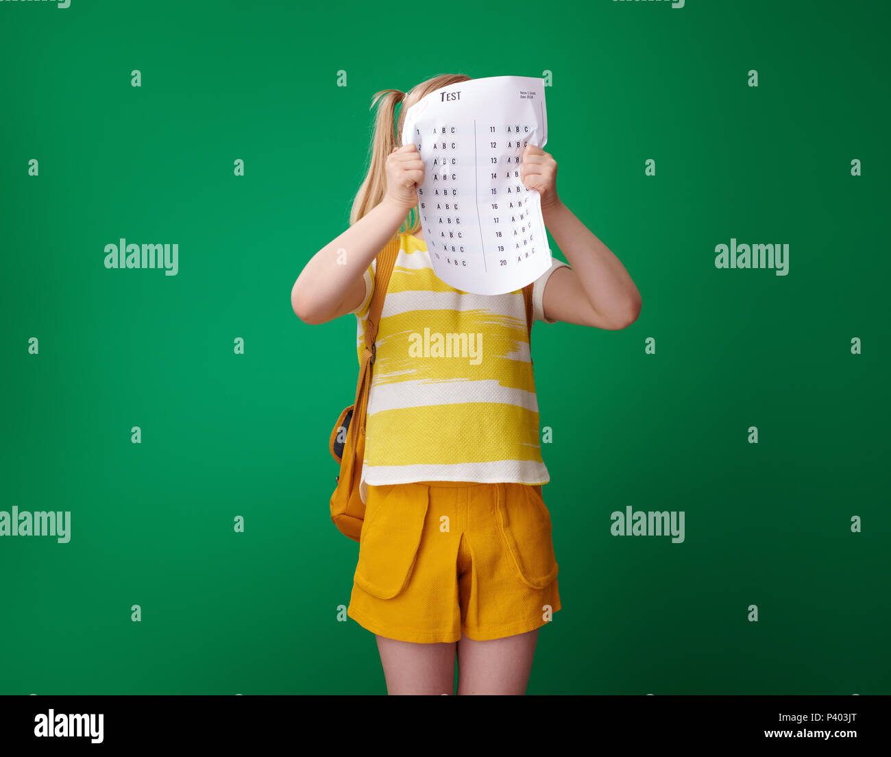 Scuola arrabbiato ragazza con zaino si nasconde dietro una cattiva prova di grado isolato su sfondo verde Foto Stock