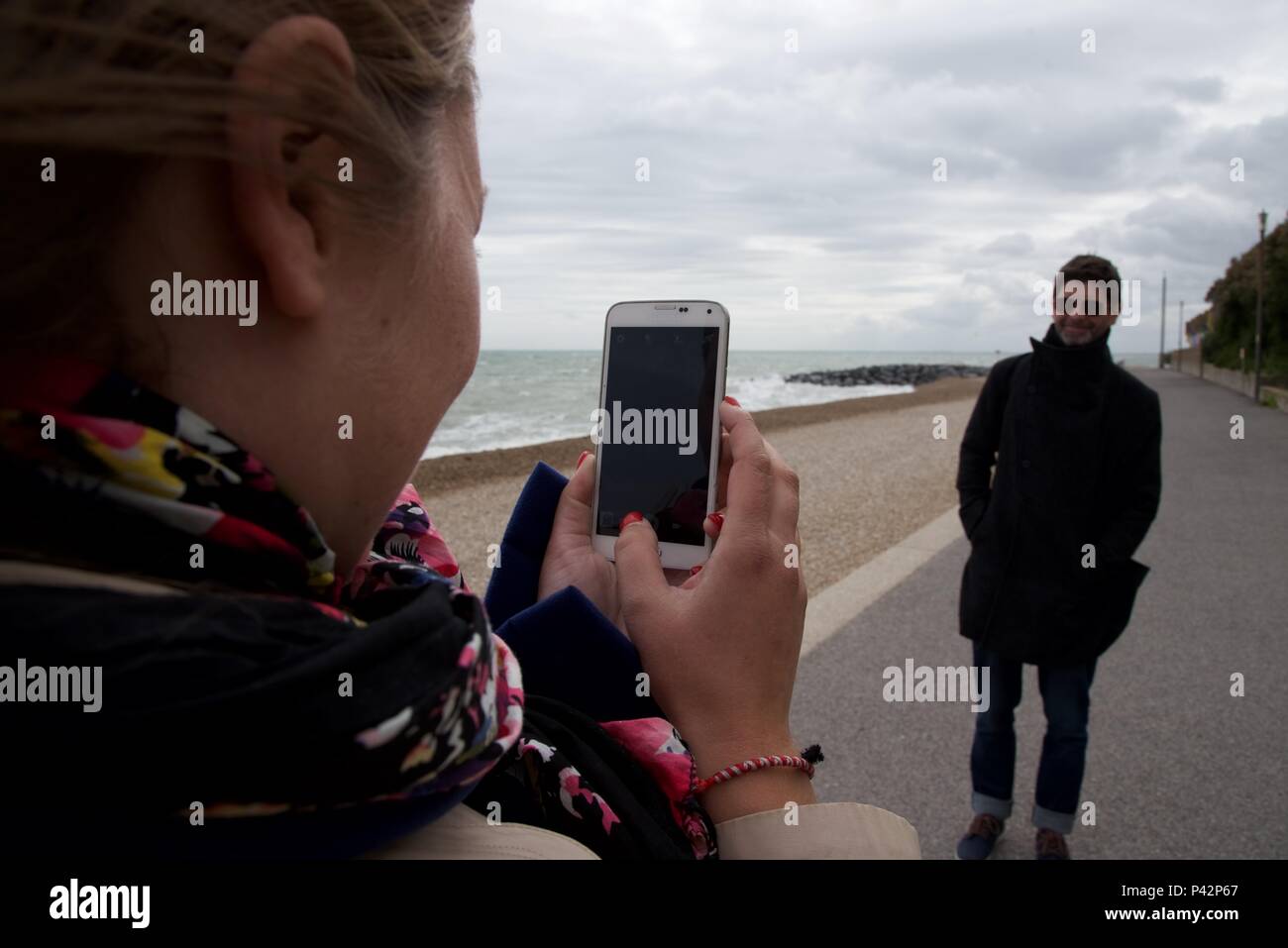 Giovane: femmina di scattare una foto del suo partner maschile sul fronte mare sullo smartphone Foto Stock