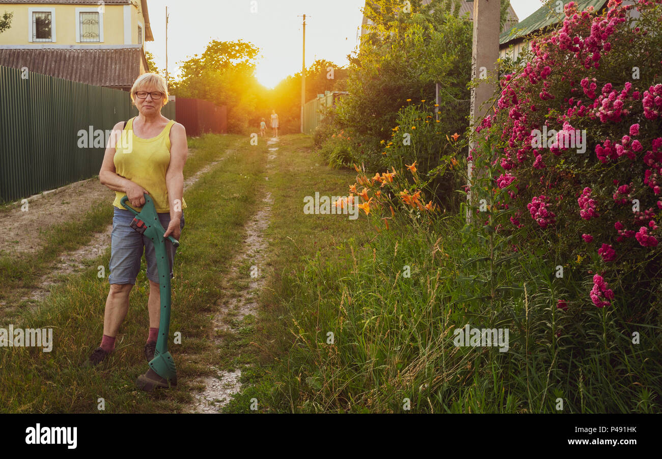 Caucasian donna di mezza età la falciatura di erba sulla strada di fronte a casa rurale con un trimmer Foto Stock