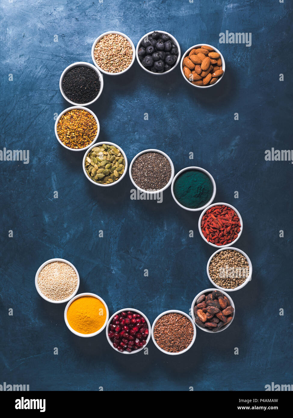 Al di sopra di vista di vari superfoods in piccolo vaso in forma S lettera su sfondo blu scuro. Super come chia,spirulina,fava di cacao,goji, canapa, mirtillo, quinoa, polline di api,sesamo nero,curcuma. Vista superiore Foto Stock