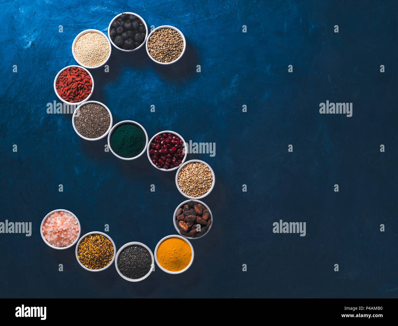 Al di sopra di vista di vari superfoods in piccolo vaso in forma S lettera su sfondo blu scuro. Super come chia,spirulina,fava di cacao,goji, canapa, mirtillo, quinoa, polline di api,sesamo nero,curcuma. Vista superiore Foto Stock