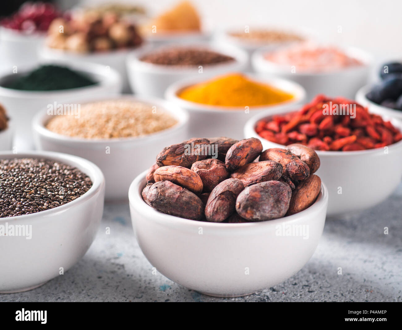 Materie fava di cacao in piccola ciotola bianco e altri superfoods sullo sfondo. Messa a fuoco selettiva. Superfoods diversi ingredienti. Concetto e illustrazione per la super e detox food Foto Stock