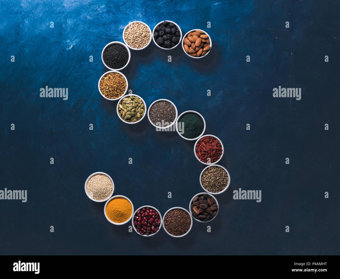 Al di sopra di vista di vari superfoods in piccolo vaso in forma S lettera su sfondo blu scuro. Super come chia,spirulina,fava di cacao,goji, canapa, mirtillo, quinoa, polline di api,sesamo nero,curcuma. Vista superiore Foto Stock