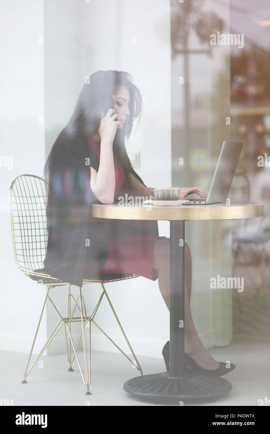 Piuttosto giovane donna parla al telefono cellulare seduti vicino al tavolo con computer portatile in cafe per interni Foto Stock