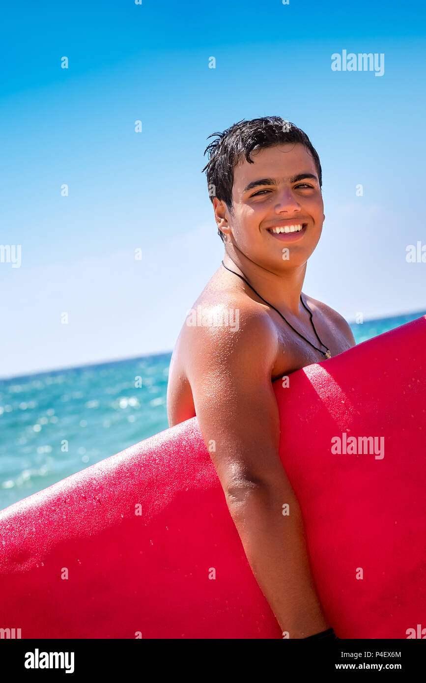 Closep ritratto di un felice allegro surfboarder rosso con la tavola da surf in piedi sopra il cielo blu di sfondo, godendo il suo hobby, uno stile di vita attivo della gioventù Foto Stock