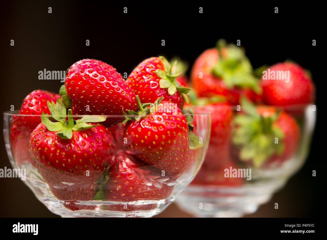 Primo piano di due piatti da dessert in vetro, pieni di fragole fresche e succose con foglie e steli, sfondo nero. Ciotola di frutta isolata all'interno. Foto Stock