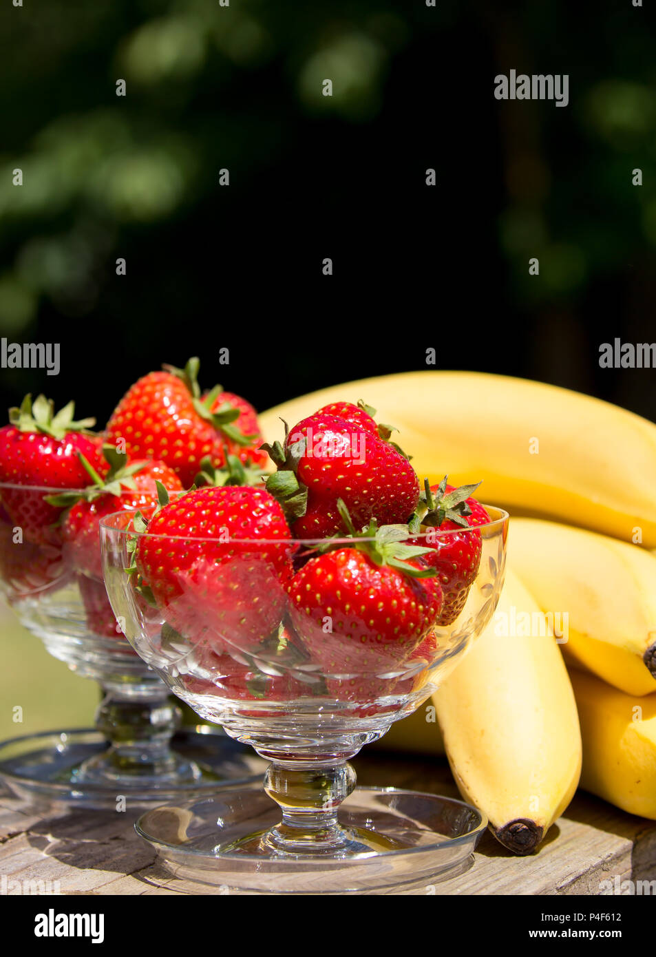 Primo piano fragole fresche in piatti da dessert in vetro e mazzo di banane all'aperto, soleggiato giorno d'estate. Godetevi le vacanze estive nel Regno Unito in giardino. Staycation. Foto Stock
