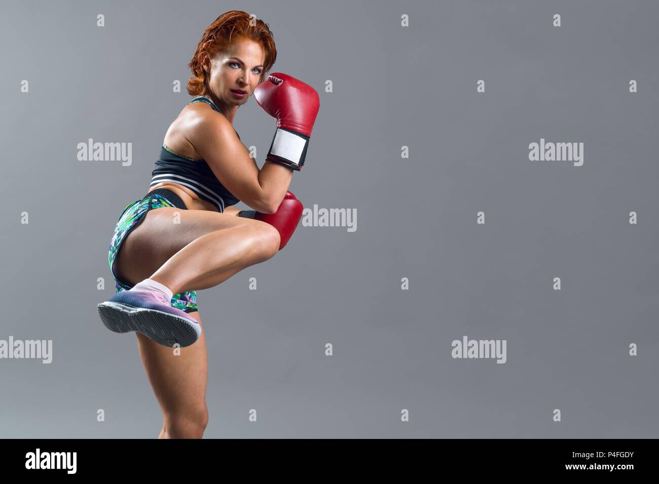 Athletic donna matura boxer con Guanti rossi in abbigliamento sportivo, guardia di sé e sempre pronti ad attaccare. Grigio di sfondo per studio, spazio di copia Foto Stock