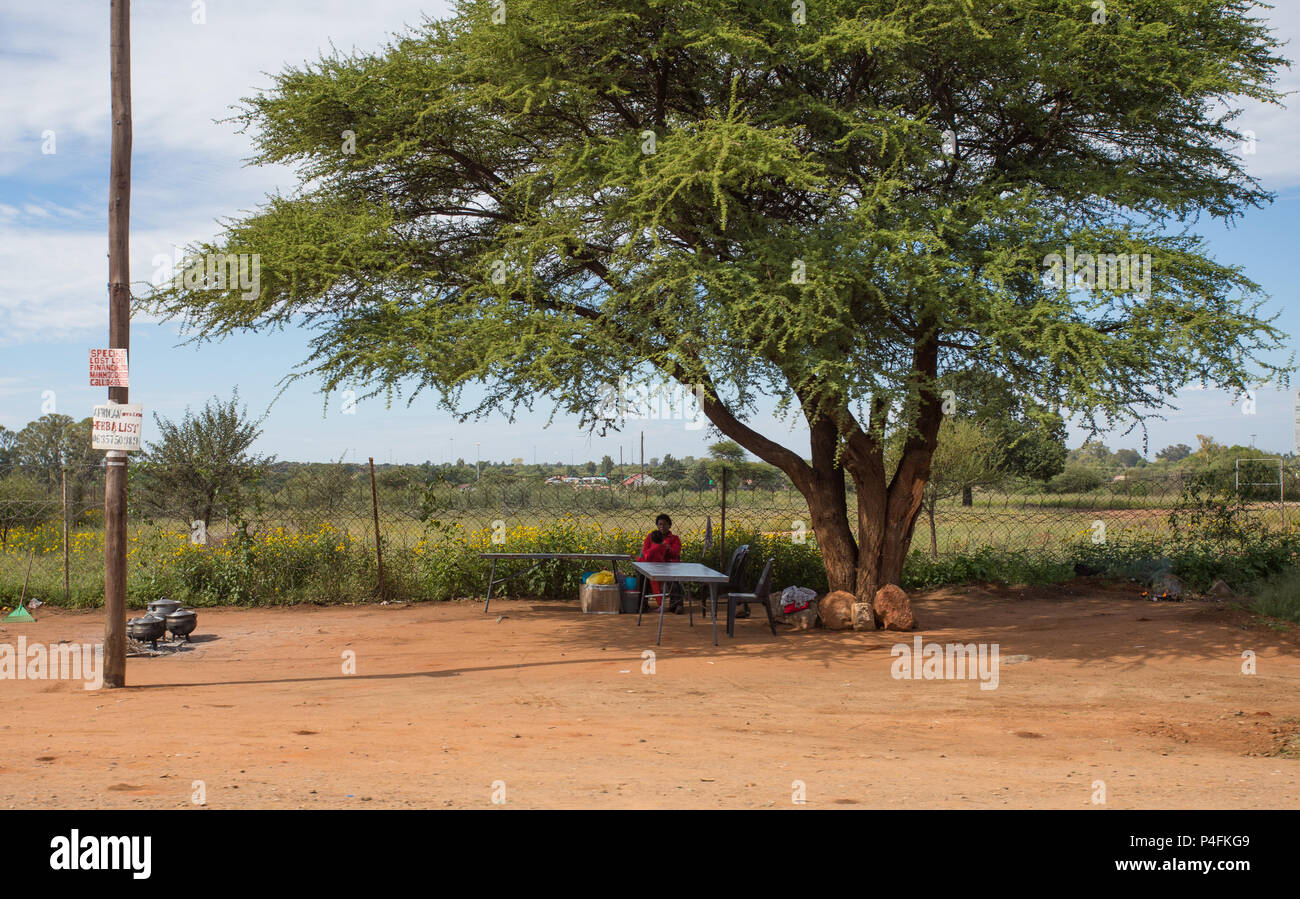 Scena rurale di una femmina africana e di un bambino seduto ad un tavolo vuoto sotto un albero di Thorn di cammello (Acacia) al lato della strada in Sudafrica Foto Stock