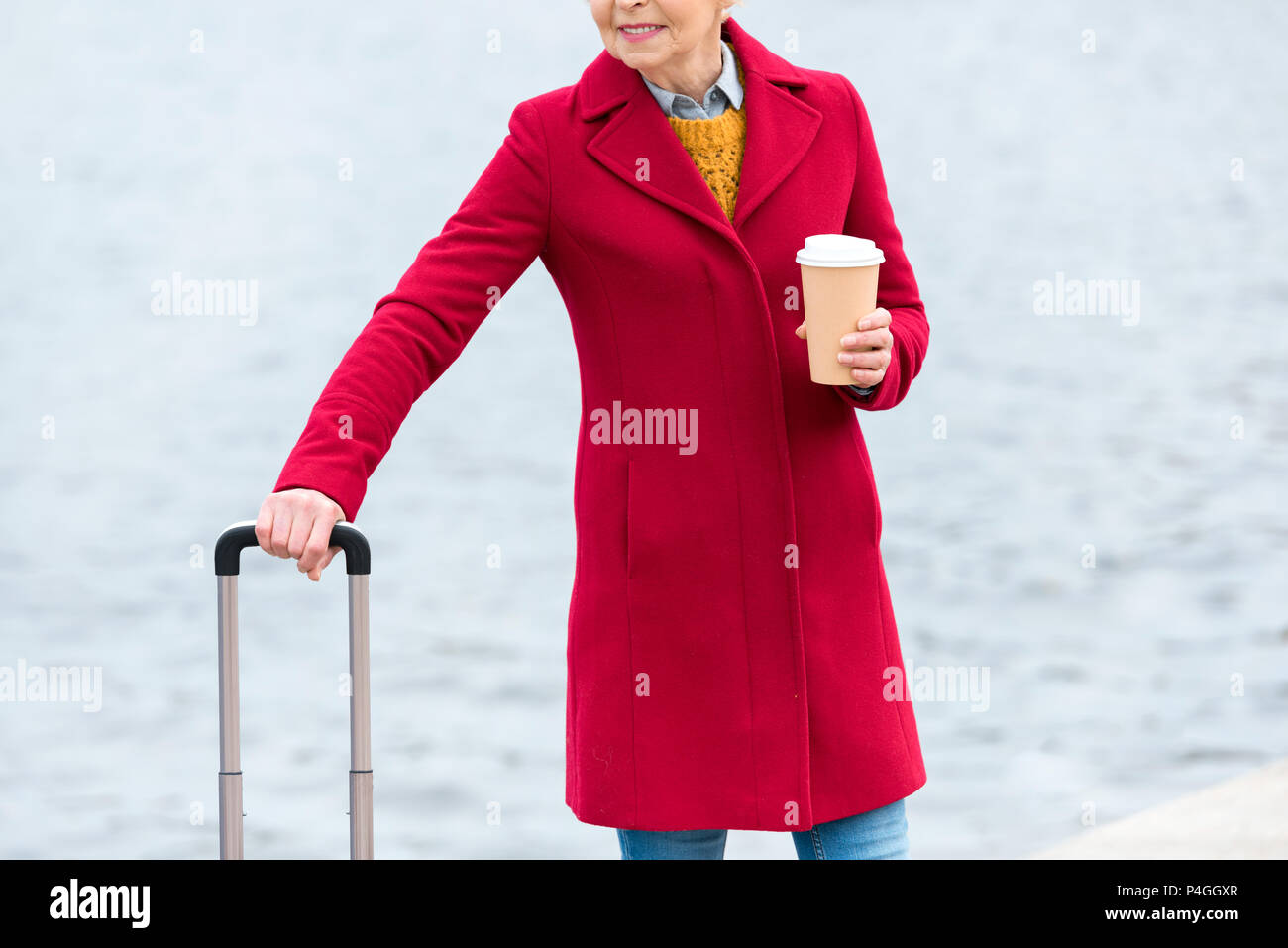 Vista ritagliata della senior woman in red coat con caffè di andare e di bagagli sul quay Foto Stock