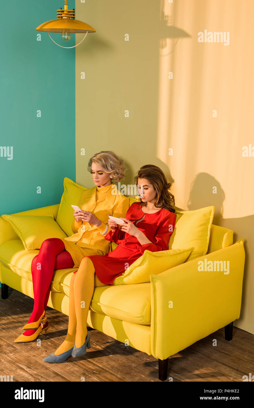 In stile retrò donne utilizzando gli smartphone mentre si riposa sul divano giallo, casa di bambola concept Foto Stock