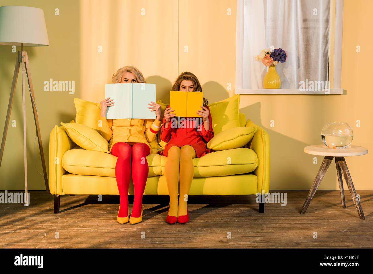 Oscurata vista delle donne in abbigliamento retrò con libri seduti sul divano giallo a luminoso appartamento, casa di bambola concept Foto Stock