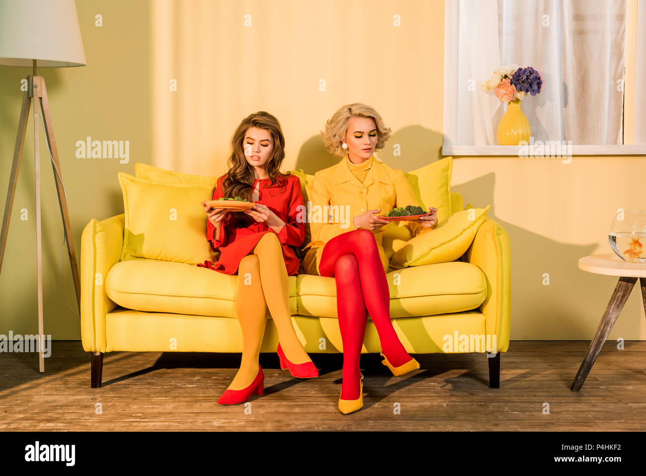 Le belle donne in abbigliamento retrò con verdure su piastre seduto sul divano giallo a camera luminosa, casa di bambola concept Foto Stock