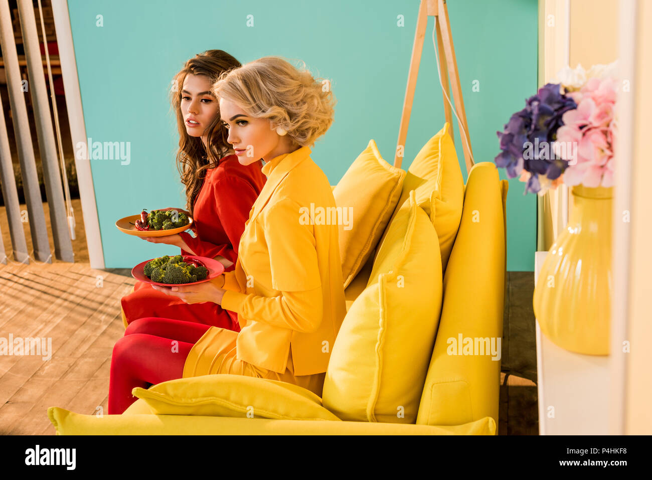 Le belle donne in abbigliamento retrò con verdure su piastre seduto sul divano giallo a camera colorata, casa di bambola concept Foto Stock