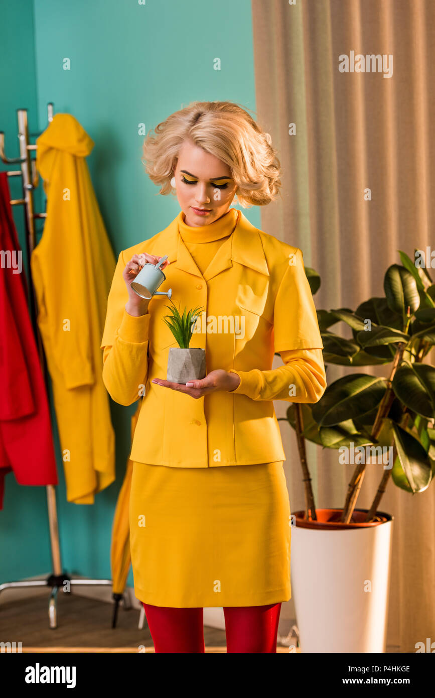 Bella vecchia donna in abito giallo di irrigazione di piante in vaso con un piccolo annaffiatoio a casa Foto Stock