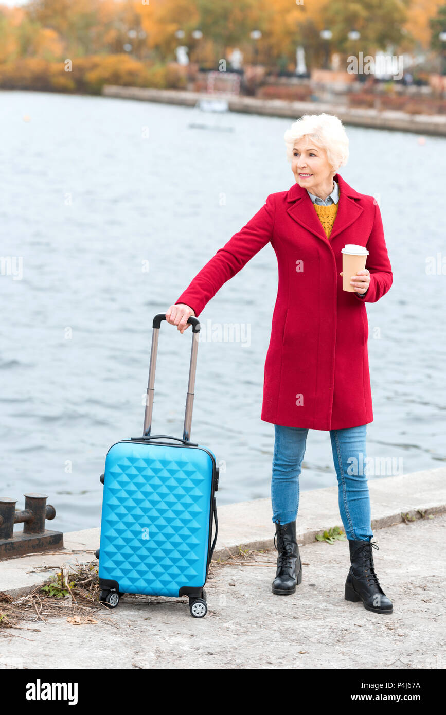 Senior woman in red coat con caffè di andare con la valigia in banchina Foto Stock