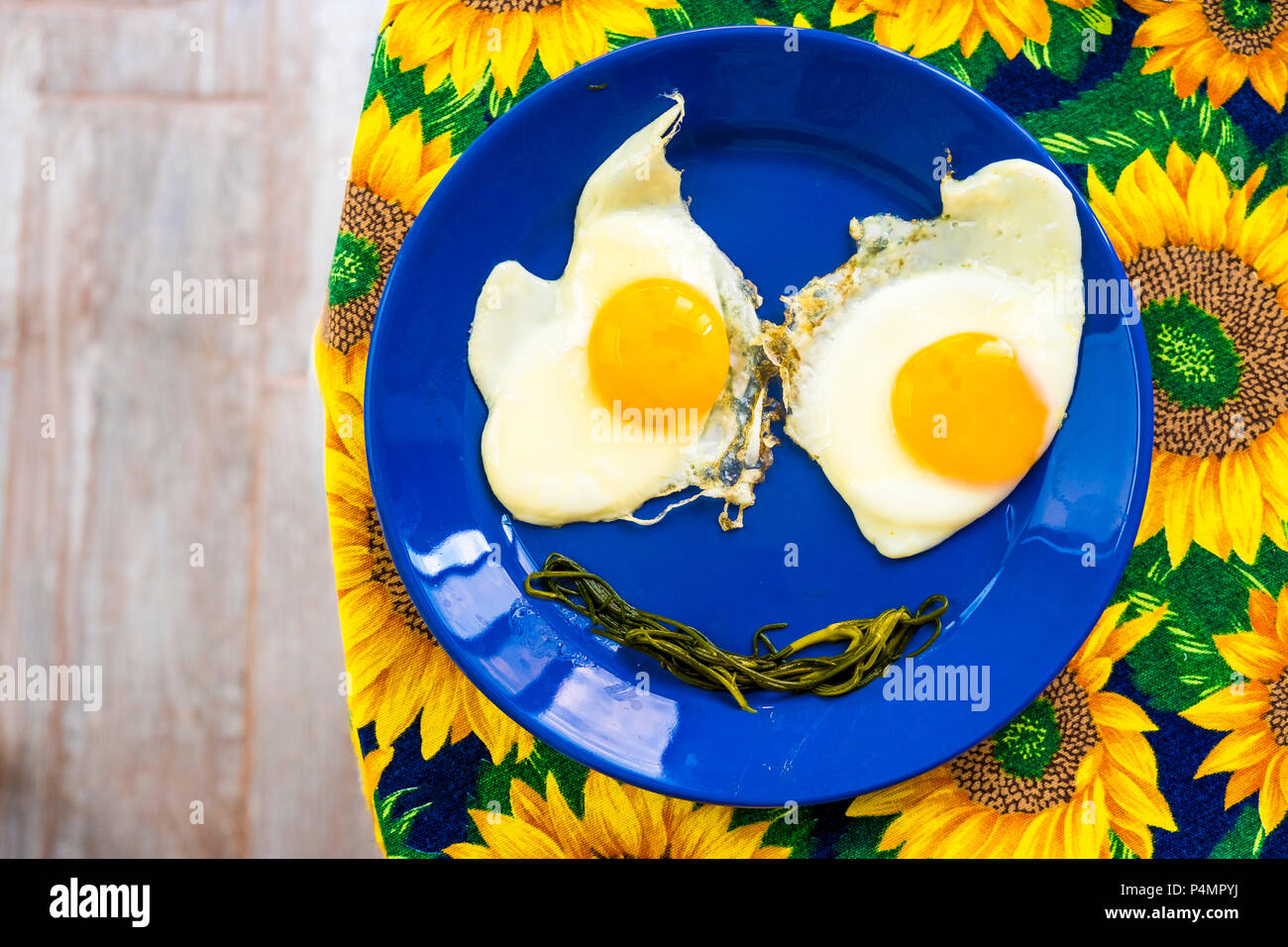 Presente la piastra di alimentare - funny facce alimentare sorriso uova fritte occhi bocca vegetale sul piatto blu giallo dei girasoli stoffa Foto Stock