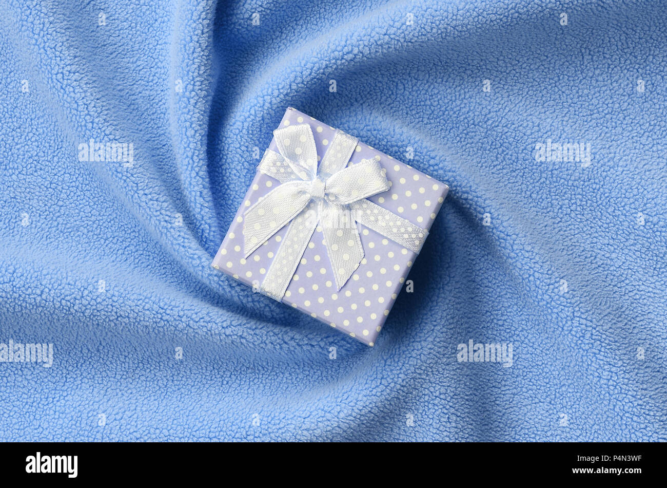 Un piccolo regalo in blu con un piccolo inchino giace su una coperta di soffice e peloso luce blu in tessuto di pile con un sacco di pieghe in rilievo. Imballaggio per un regalo Foto Stock