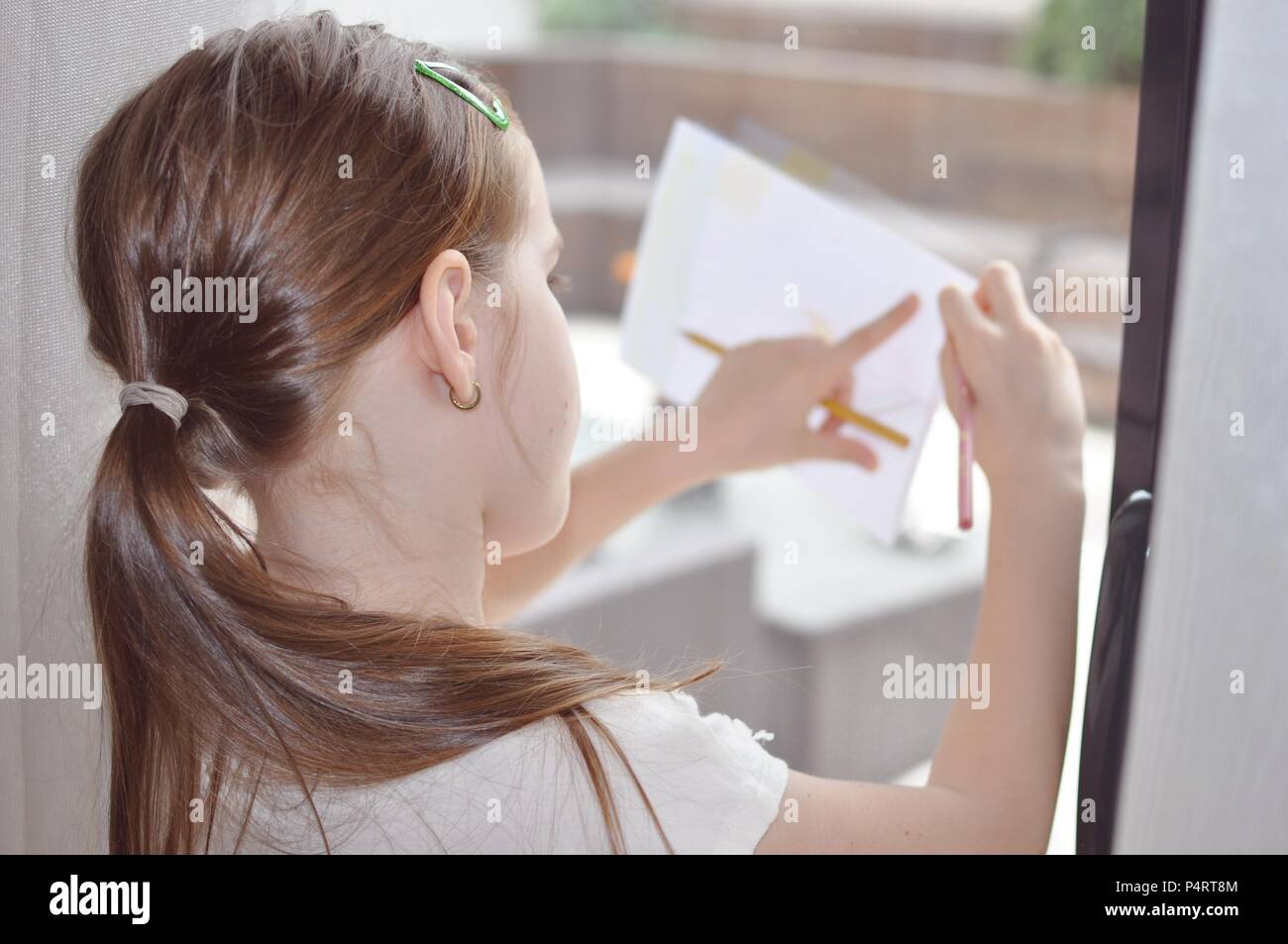 Bambino, ragazza 7-9, capelli biondi, disegno su un foglio bianco di carta supportato sul vetro di messa a fuoco selettiva, ritratto indietro Foto Stock
