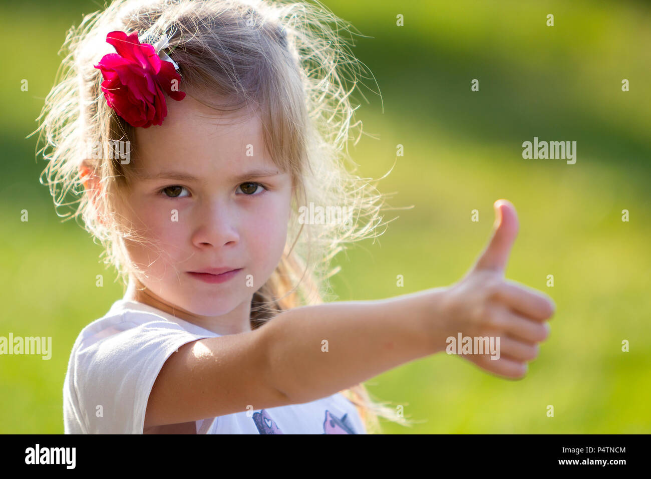 Ritratto di carino piccolo bionda ragazza gravi in T-shirt bianco con rosa rossa nei capelli che mostra il pollice-fino al gesto fotocamera su sfocato bright green estate bac Foto Stock