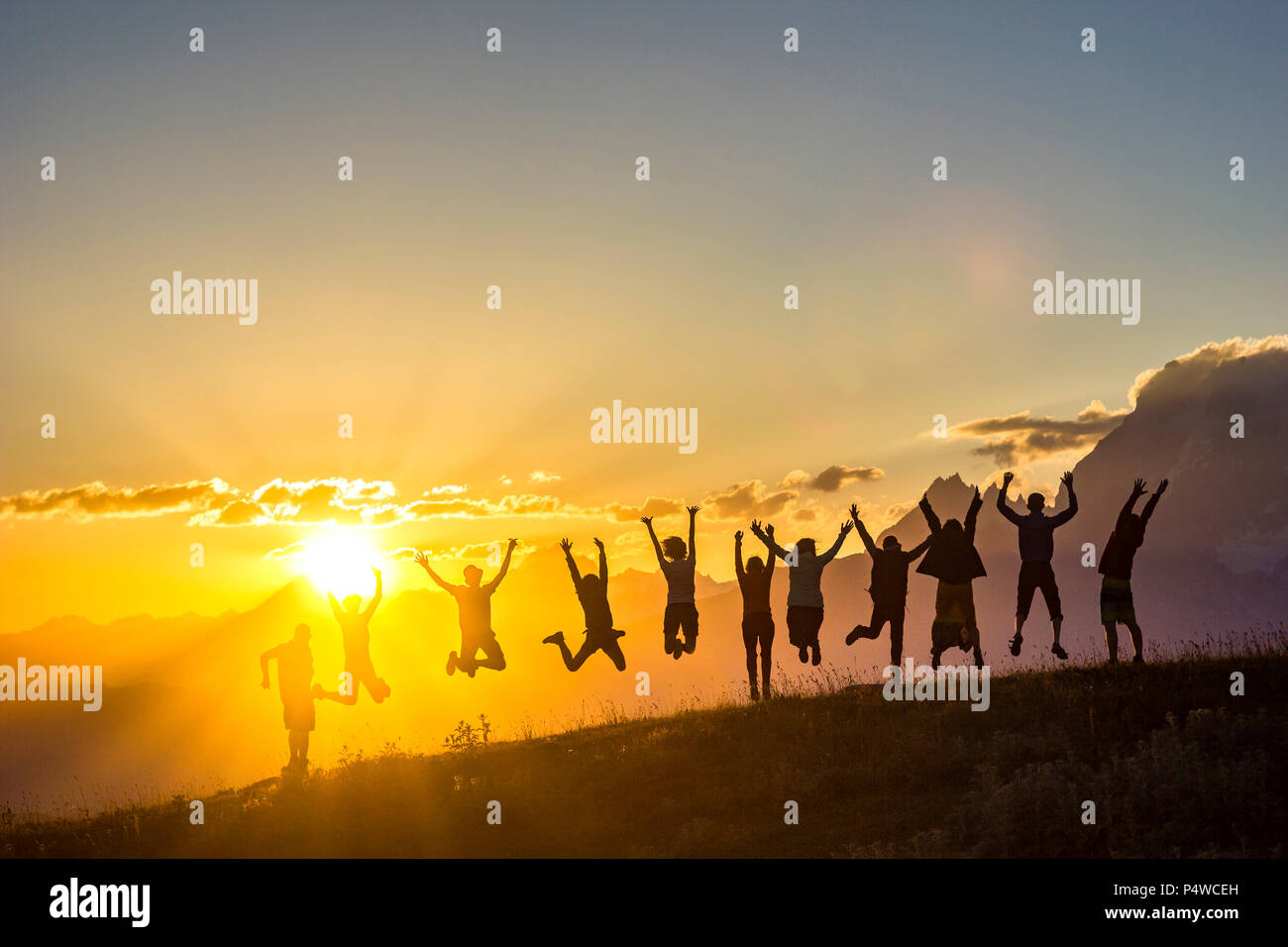 Un gruppo di persone con le mani in alto saltando su erba in montagne al tramonto Foto Stock