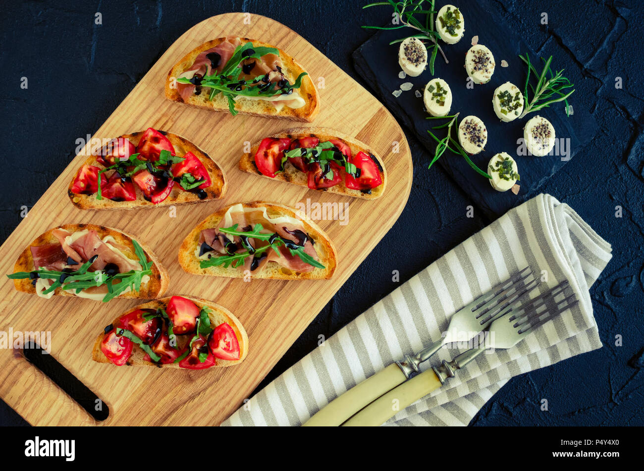 Italian bruschetta con pomodori, Prosciutto di Parma, rucola e balsamico salsa glasse sul tagliere e mini antipasti di formaggio su backgroun scuro Foto Stock