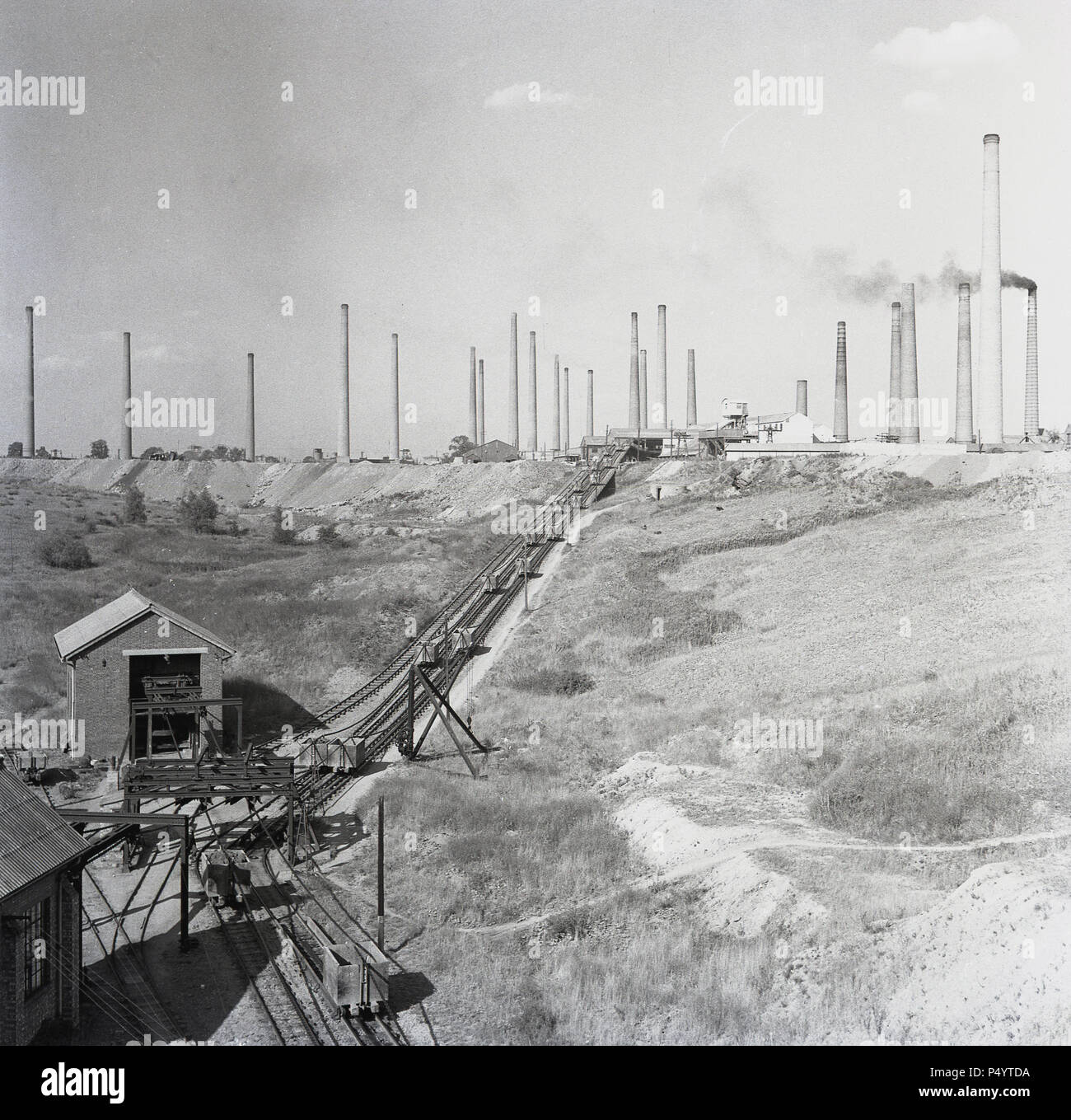 Anni '50, su una cava o una fossa di mattoni in disuso presso la London Brick Company a Stewartby, Bedford, Inghilterra, UK, vediamo le piste ferroviarie utilizzate per trasportare la roccia minata e in lontananza i numerosi camini dei muratori, che alla sua vetta erano tanti 162. Dopo la guerra questa società britannica era il più grande produttore di mattoni del mondo, con l'estrazione di "Oxford Clay", la pietra naturale organica o la roccia, la materia prima per il famoso London Brick. A Stewartby, costruì uno dei pochi 'Model Villages' del XX secolo, fornendo alloggi, sport e strutture sociali per la sua grande forza lavoro industriale. Foto Stock