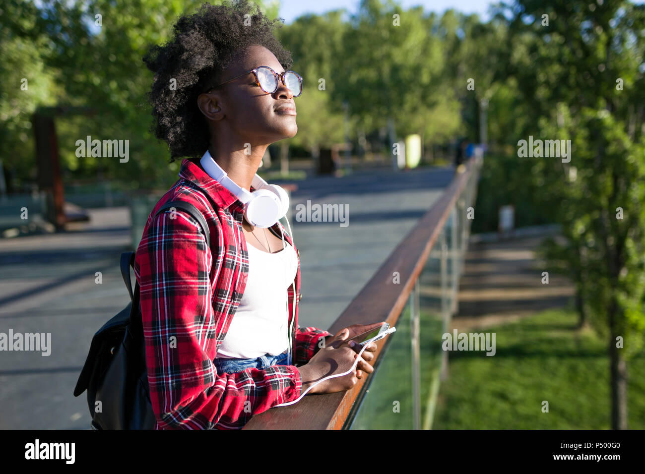 Giovane donna con le cuffie e il telefono cellulare in piedi su un ponte godendo la luce solare Foto Stock