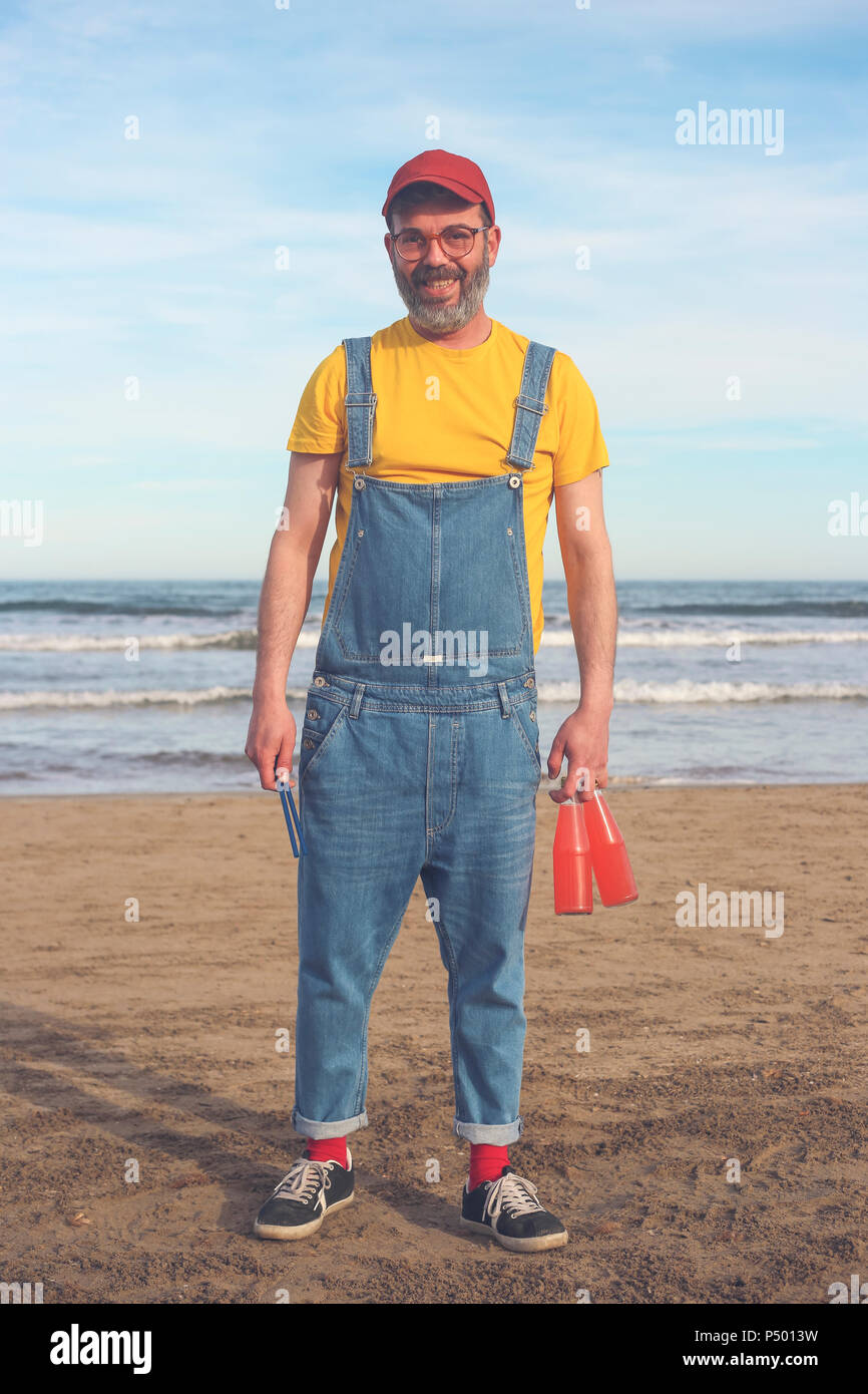 Ritratto di uomo sorridente in salopette da permanente sulla spiaggia tenendo le bottiglie di bevande analcoliche Foto Stock