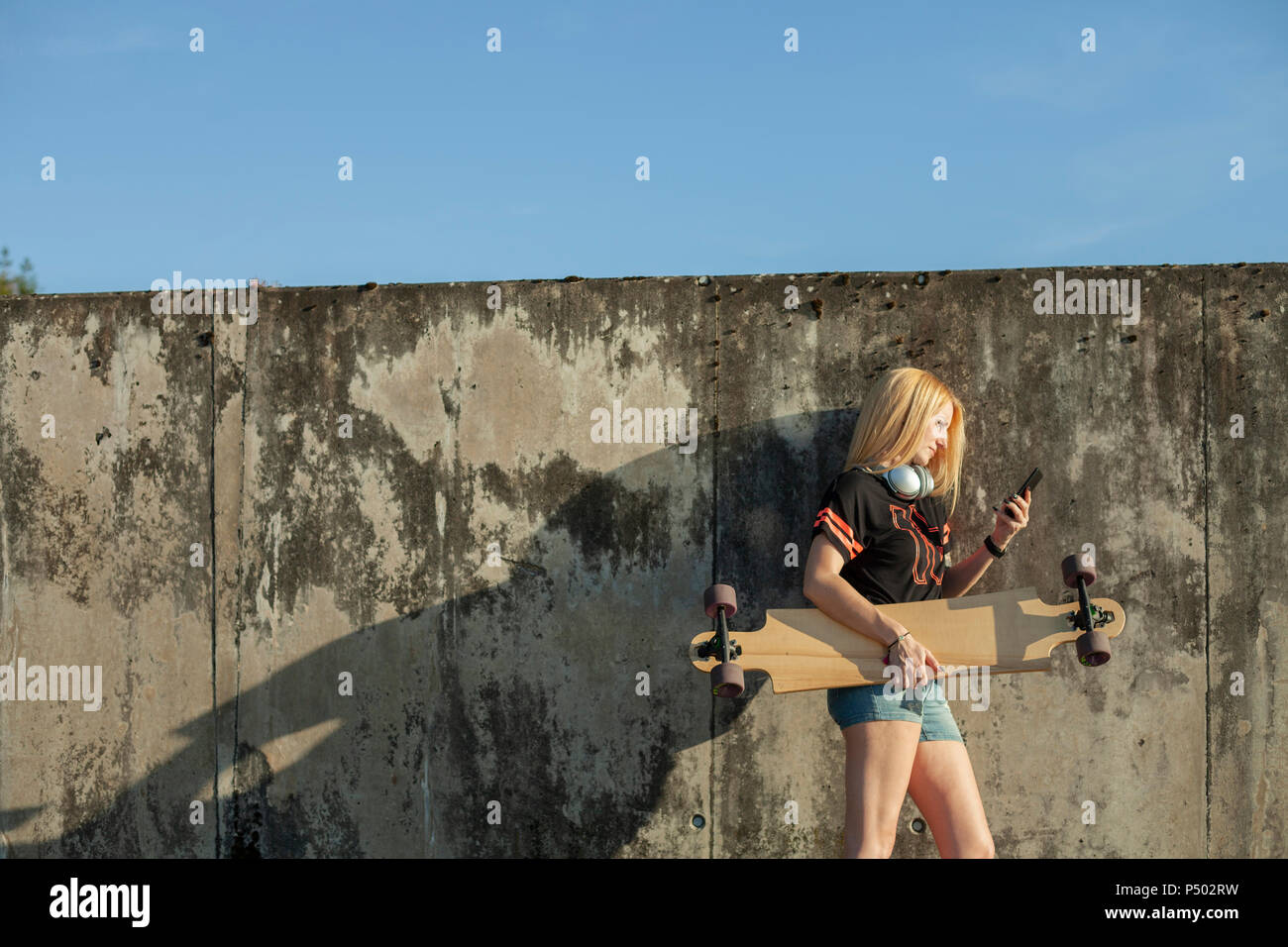 Donna bionda con longboard in piedi nella parte anteriore della parete guardando smartphone Foto Stock