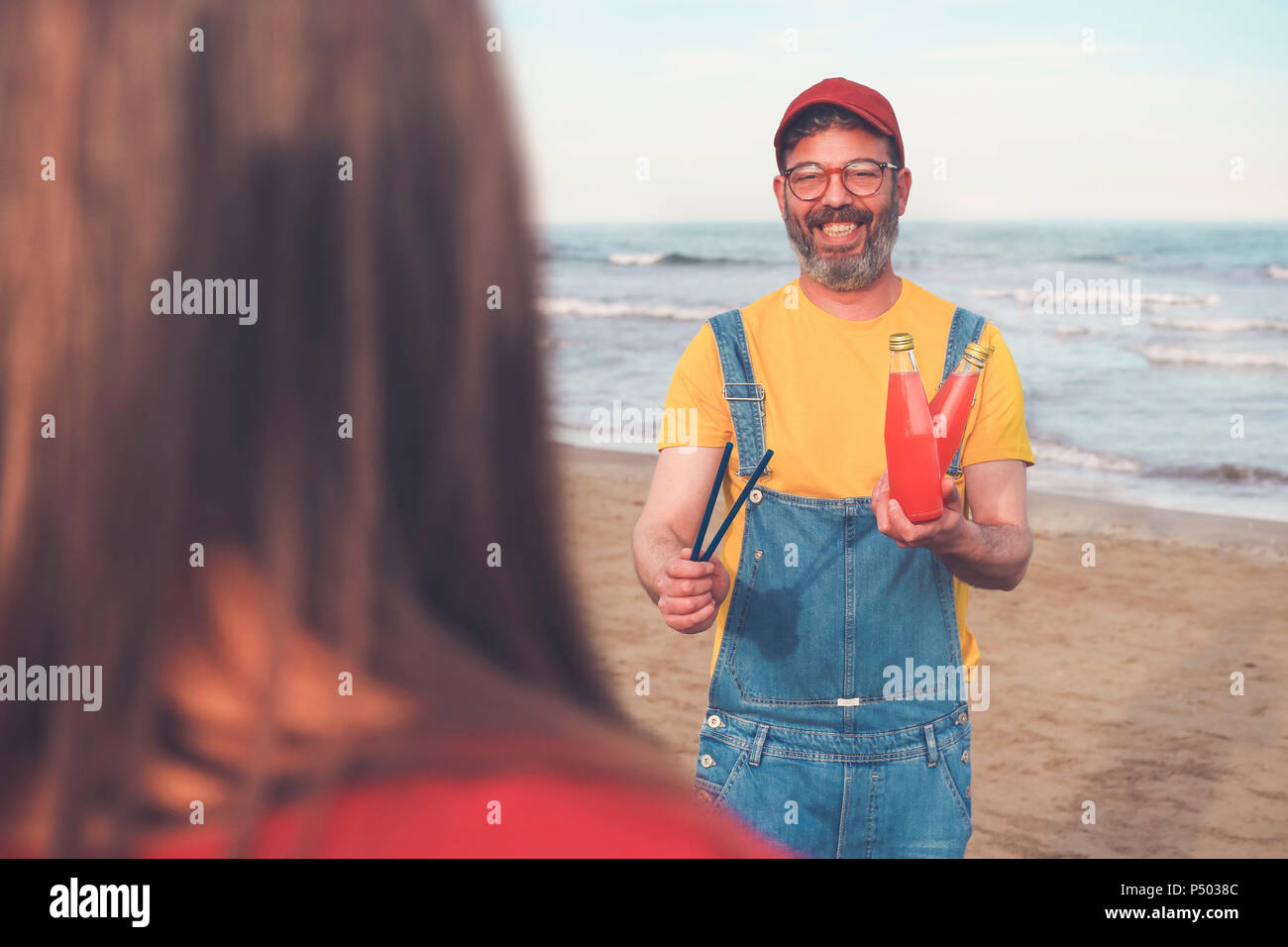 Uomo felice in salopette da sulla spiaggia che offre soft drink per donna Foto Stock