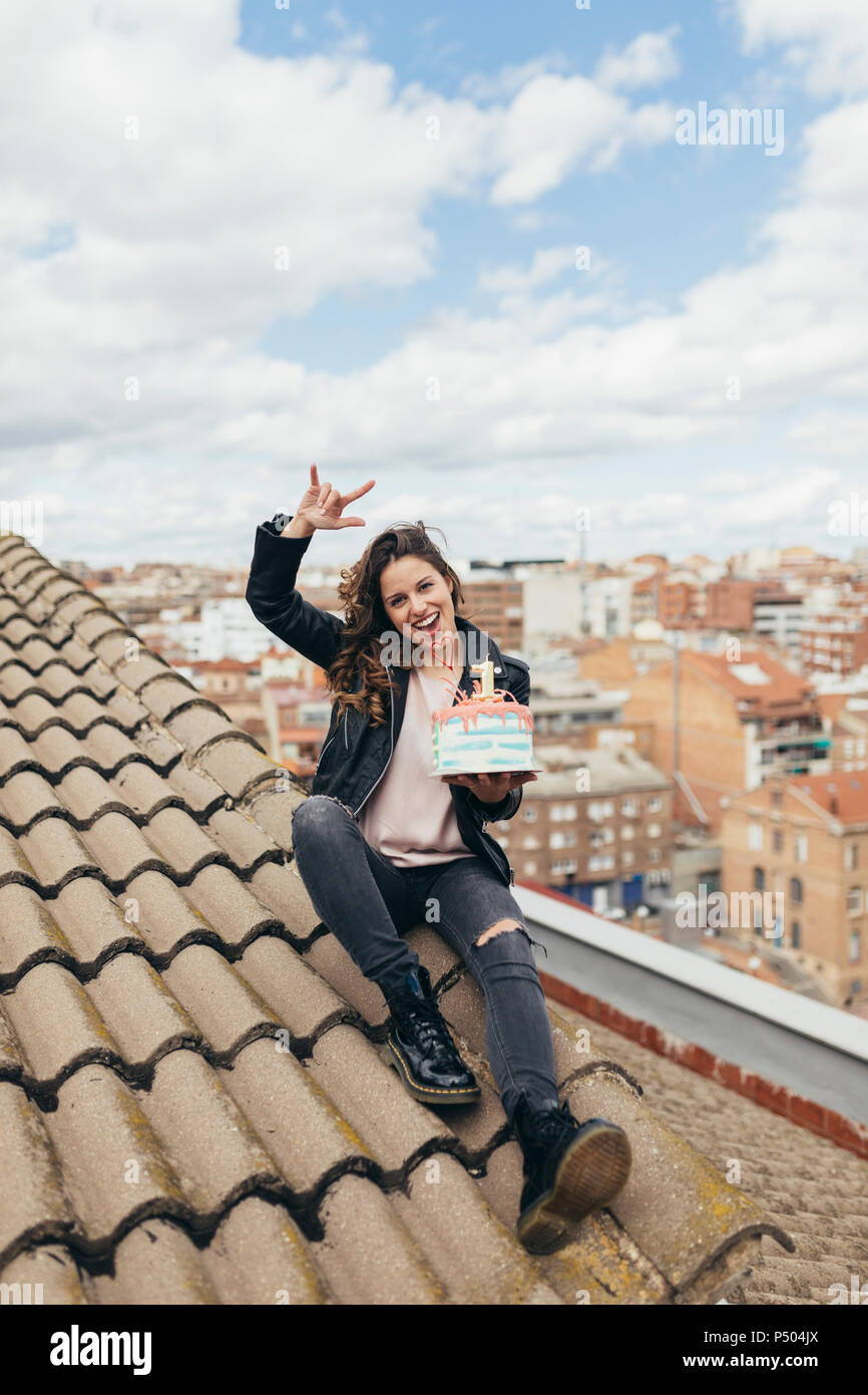 Ridendo donna seduta sul tetto con torta di compleanno che mostra Rock And Roll segno Foto Stock