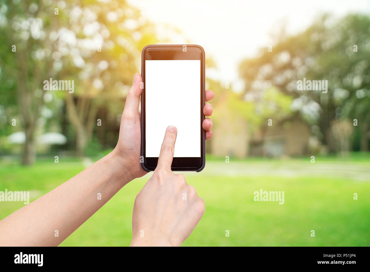 Mano che tiene il telefono con defocalizzata bokeh di fondo e la sfocatura sullo sfondo di un giardino di alberi nella luce solare. Foto Stock
