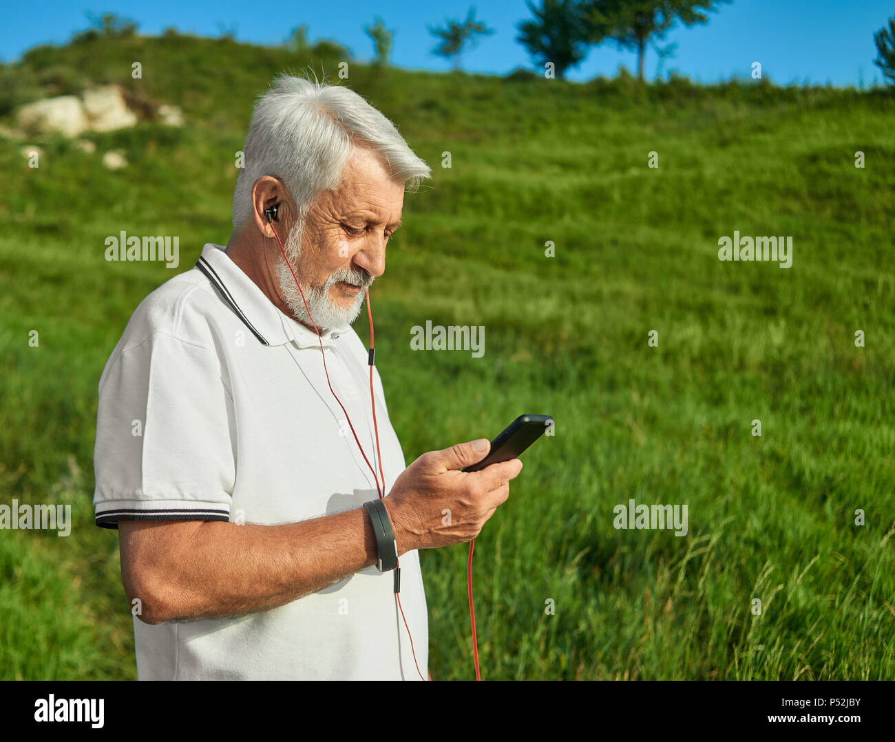 Il vecchio uomo che guarda sul suo cellulare all'esterno. Indossando il bianco classico Polo shirt con blu scuro strisce, sportwatch, ascoltare musica con le cuffie. Stile di vita sportiva. Una buona sensazione positiva. Foto Stock