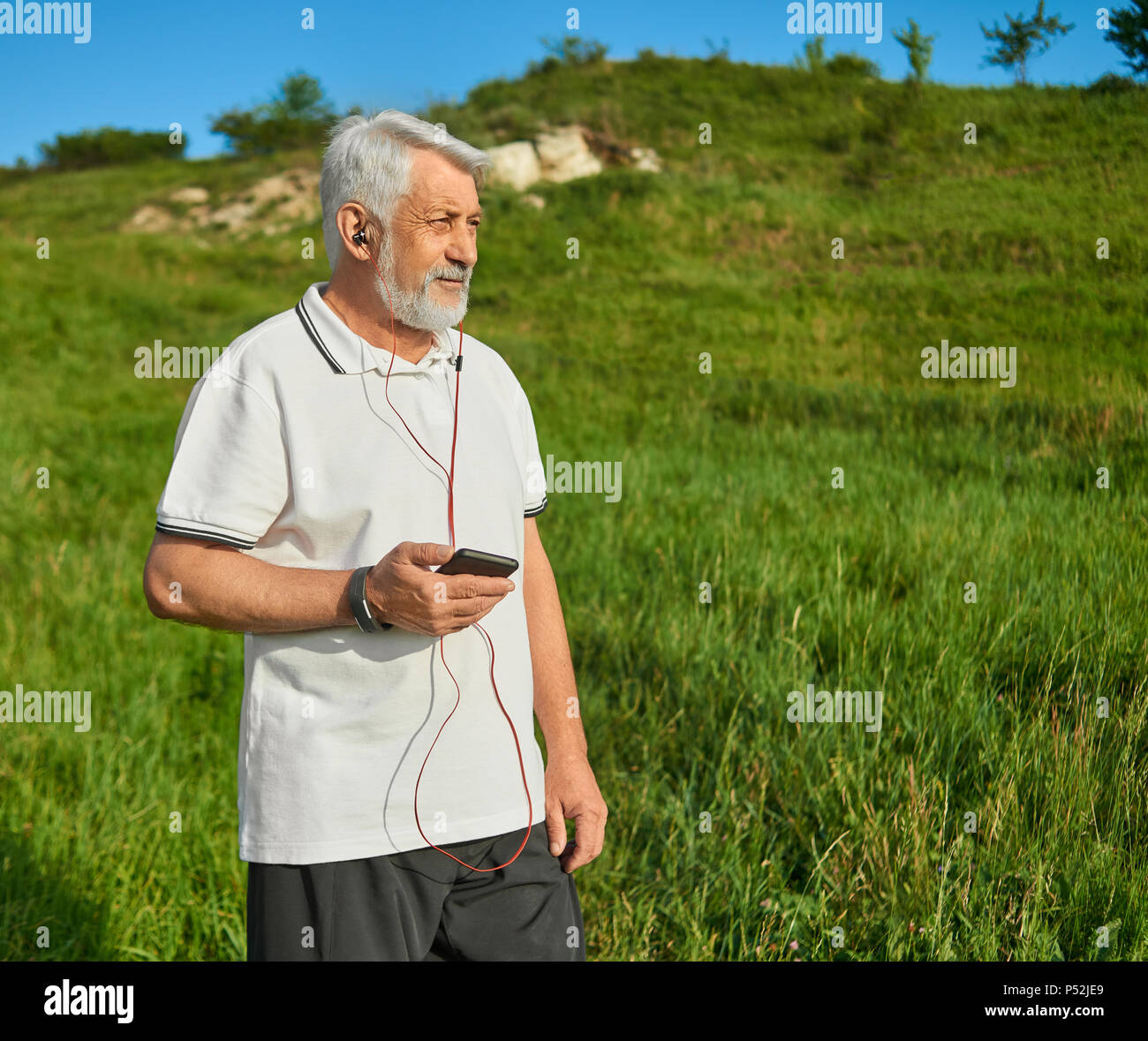 Il vecchio uomo tenere il cellulare acceso sulla campagna. Ascolto di musica con cuffie rosso. Indossando il bianco classico Polo shirt scuro con strisce blu,pantaloni neri, sneakers. Stile di vita sportiva. Foto Stock