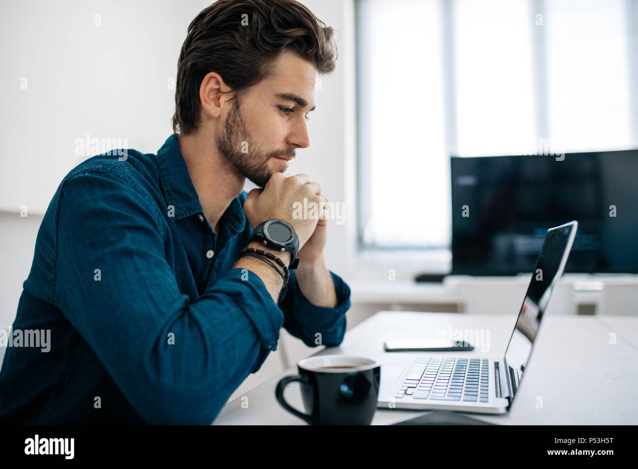 Software Developer seduti nella parte anteriore del computer e lavorare in ufficio. Uomo che guarda un notebook seduto con il suo mento appoggiato sulle mani con una tazza di caffè Foto Stock