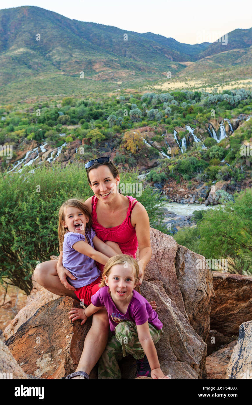 Madre di due ragazze seduta sulle rocce nel verde paesaggio, dietro Epupa Falls, Kunene, regione di Kunene, Namibia Foto Stock