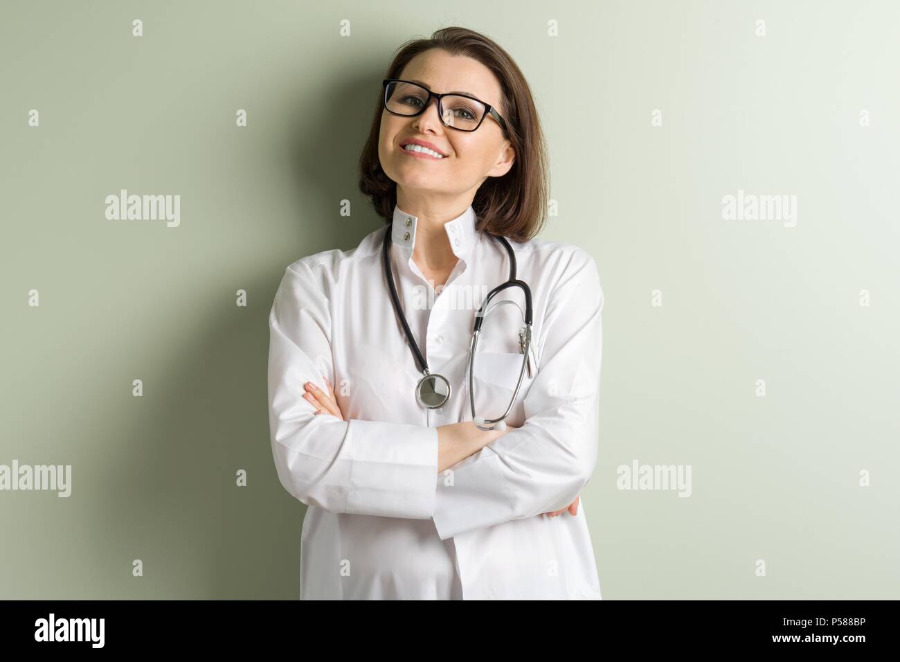 Positivo donna di mezza età medico, indossando occhiali con bracci incrociati. Sfondo verde parete in clinica Foto Stock