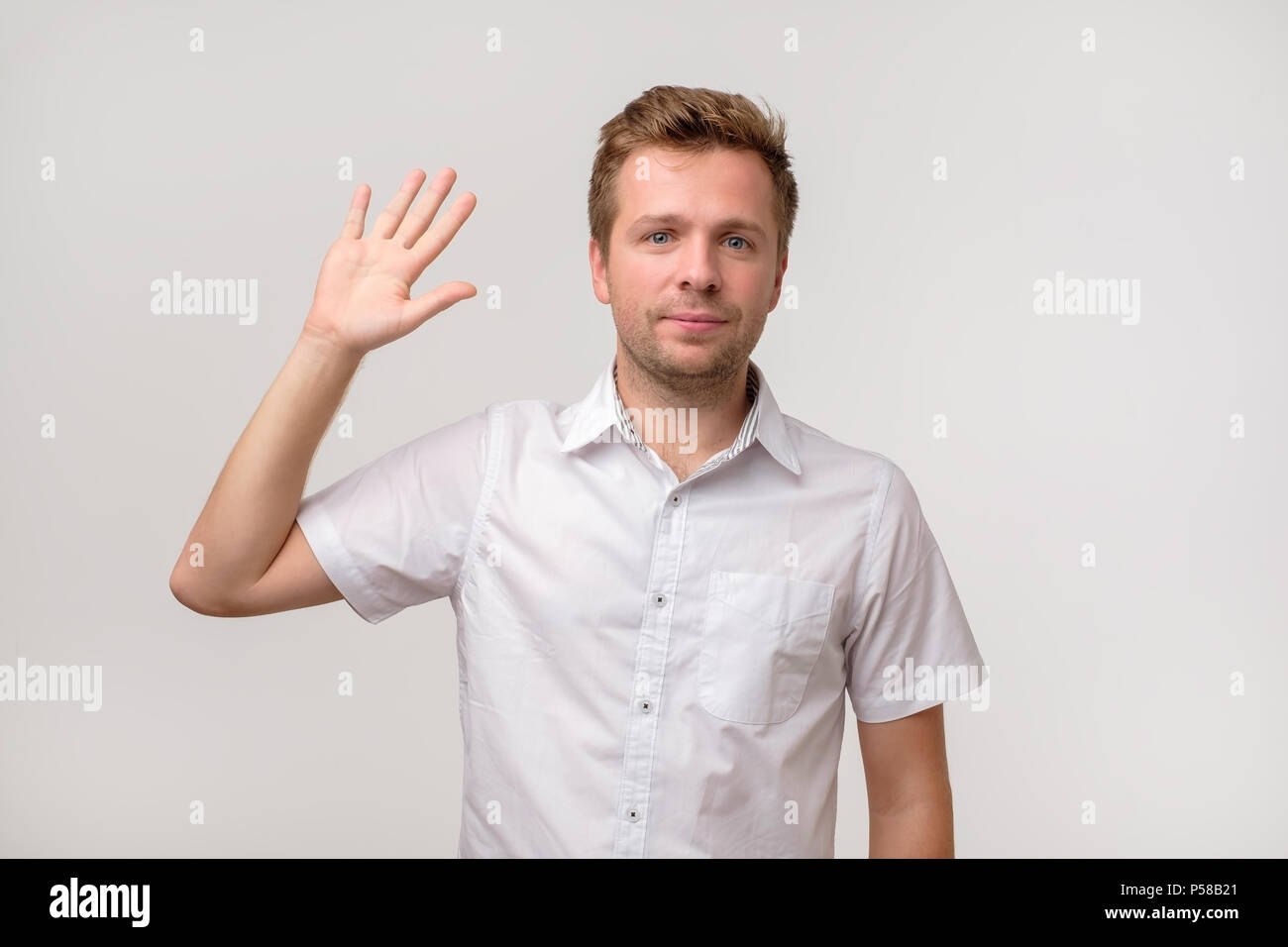 Bello attraente uomo europeo rinuncia a mano nel gesto ciao mentre sorridente allegramente. Egli è felice di dare il benvenuto ai suoi amici o ospiti. Per conoscere Foto Stock