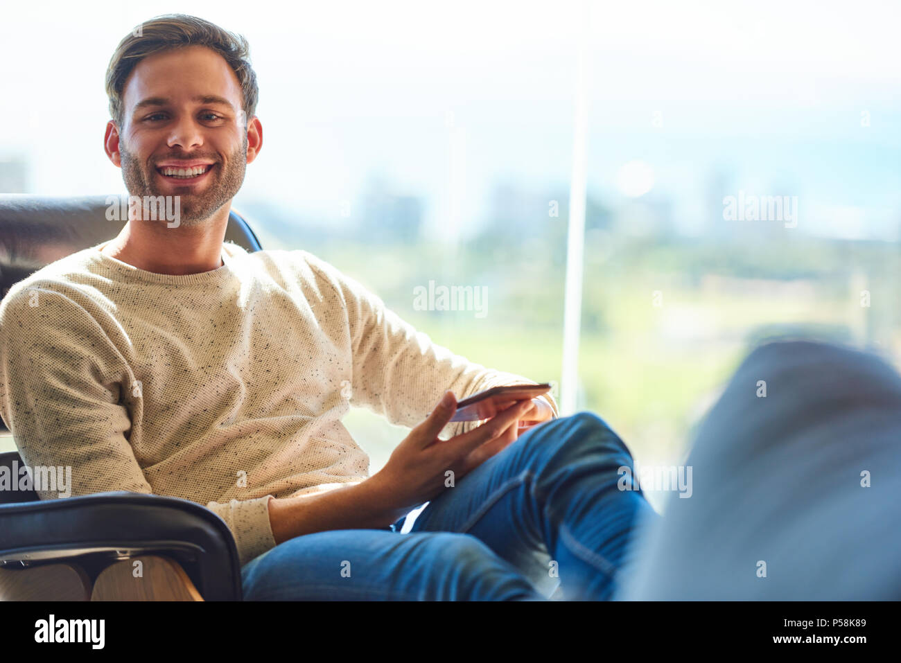 Uomo caucasico seduto su un divano moderno accanto a grandi finestre di vetro con una vista stupefacente dietro di lui come egli sorride alla telecamera con il suo telefono cellulare ancora detenuti nelle sue mani. Foto Stock