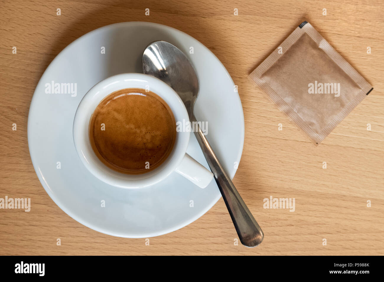 Piccoli caffè espresso italiano in bianco coppa in ceramica con cucchiaino e pacchetto di zucchero isolato luce sulla scrivania di legno dal di sopra. Foto Stock