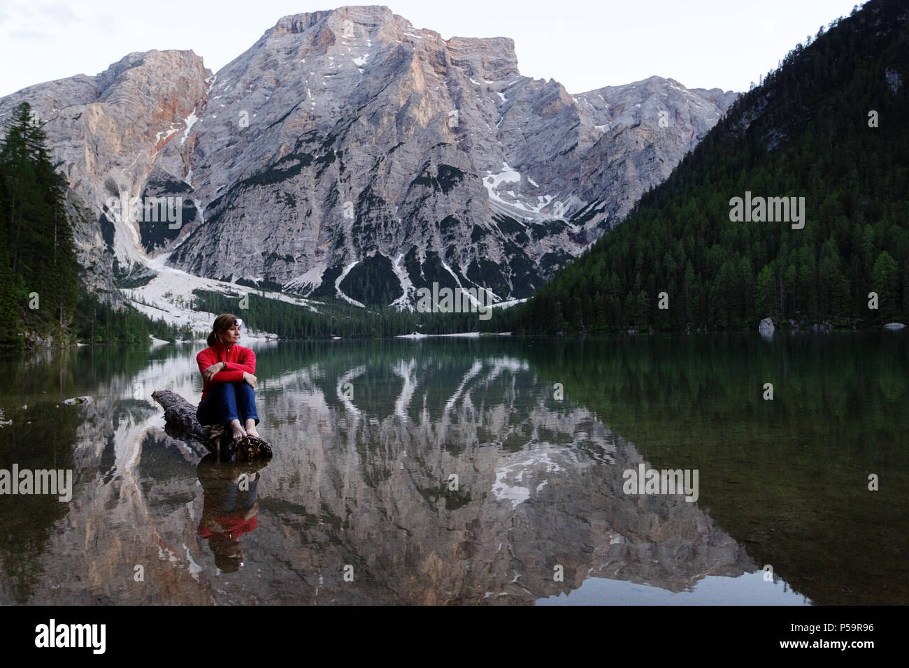 Donna che indossa un proiettore in giacca rossa seduta a piedi nudi su un registro affondata al Lago di Braies, Dolomiti, Italia Foto Stock