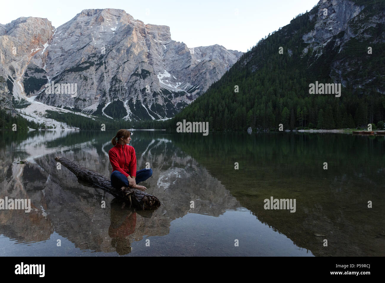 Donna che indossa un proiettore in giacca rossa seduta a piedi nudi su un registro affondata al Lago di Braies, Dolomiti, Italia Foto Stock