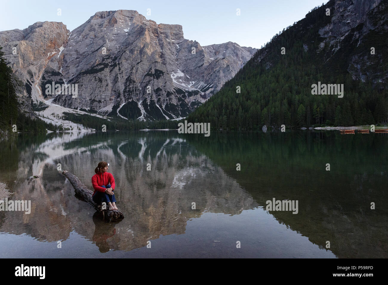Donna che indossa un proiettore in giacca rossa seduta a piedi nudi su un registro affondata al Lago di Braies, Dolomiti, Italia Foto Stock