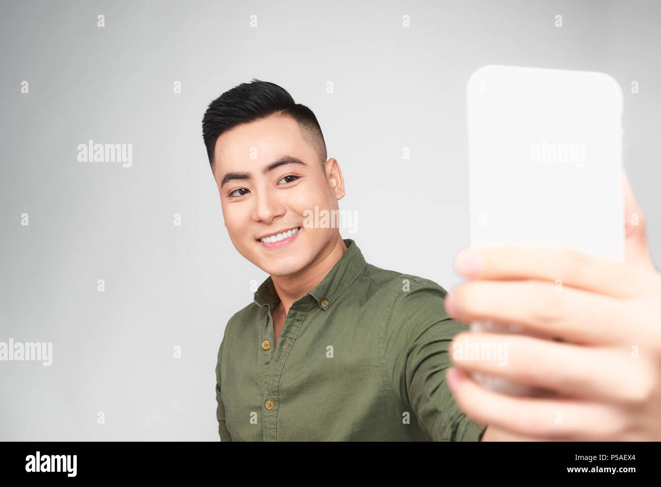 Bel ragazzo in camicia bianca si sta facendo selfie utilizzando uno smart phone, mostrando Ok segno e sorridente, su sfondo grigio Foto Stock