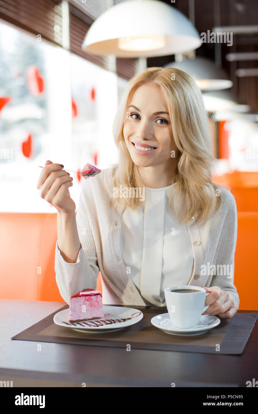 Molto bella e felice, giovane donna, sedersi al bar e mangiare il dessert con caffè o tè, ritratto verticale Foto Stock
